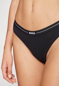 Sous-vêtements bikini noirs en tissu lisse, avec une ceinture blanche contrastante arborant le logo "BOSS", au design minimaliste et à taille basse.