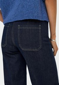 Tumši džinsu bikses ar aizmugurējām kabatām, kurām ir kontrastējoša dzeltena šuvuma apdare, apvienotas ar teksturētu zilu adītu topu. Tuvplāns no sāniem.