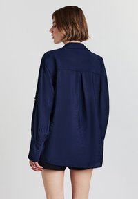 Camicetta leggera di colore blu navy con una vestibilità relax, dotata di un colletto piccolo e polsini buttoned. La texture appare leggermente stropicciata.