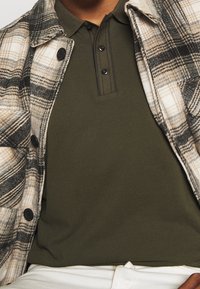 Rutig flanelljacka i kräm, svart och brun toner, lagd över en mörkgrön polotröja. Har knappar och en klassisk krage.