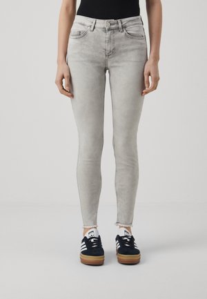 Person trägt hellgraue Skinny-Jeans mit ausgefransten Säumen und schwarze Sneakers mit weißen Streifen, steht vor einfarbigem Hintergrund.