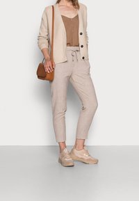 Beige vest, bruin shirt met knopen, lichtgekleurde pantalon op maat, en beige sneakers. Bruine crossbodytas, minimale patronen en texturen.