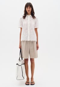 Blusa bianca in cotone con design a occhiello, maniche corte e chiusura a bottoni. Shorts beigi a pieghe, sandali neri e una borsa a tracolla bianca con bordo nero.