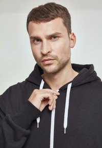 Sudadera negra con capucha y cordones blancos, que cuenta con un bolsillo tipo canguro, puños de canalé y un ajuste casual. Textura de tela suave.