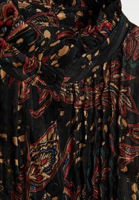Blouse noire plissée avec des motifs floraux colorés en rouge, or et bleu. Tissu texturé avec fermeture à boutons au niveau du col.
