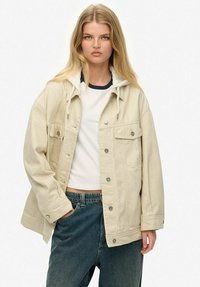 Blond kvinde iført en beige knapjakke med hætte over en hvid cropped T-shirt og blå jeans, stående med den ene hånd i lommen.