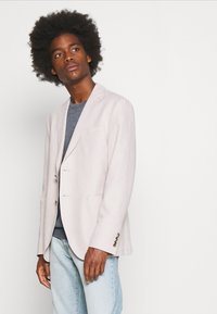 Light beige linen blazer with notch lapels, single-breasted design, and two front pockets. Paired with a gray t-shirt and light denim jeans.