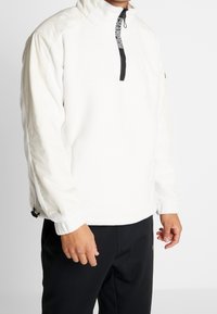 Osoba w białej polarowej bluzie z zamkiem błyskawicznym w czarnym kolorze z napisem "CHAMPION" oraz w czarnych spodniach, na jednolitym białym tle.