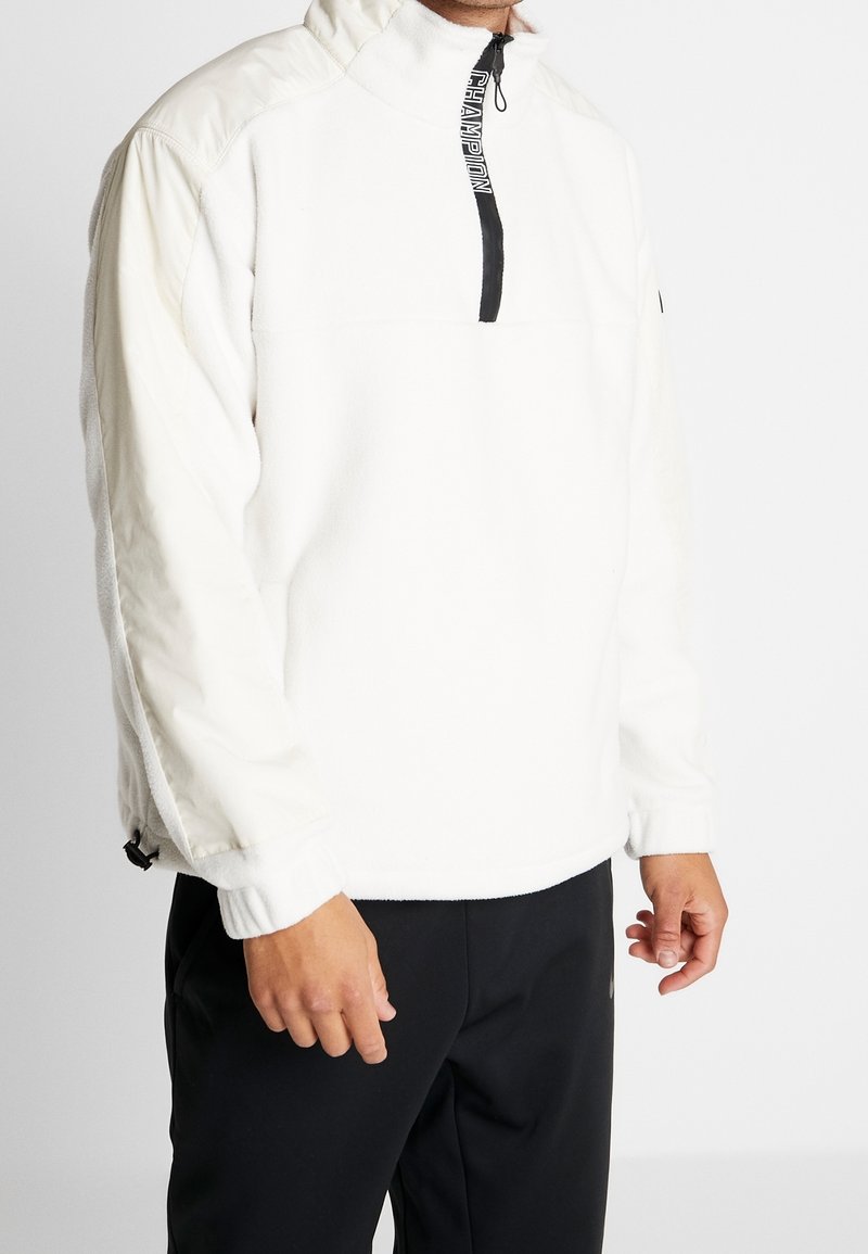 Osoba w białej polarowej bluzie z zamkiem błyskawicznym w czarnym kolorze z napisem "CHAMPION" oraz w czarnych spodniach, na jednolitym białym tle.