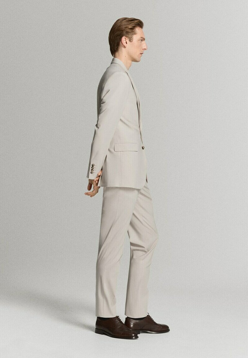 Hombre de pie con traje beige y zapatos de vestir marrones, con las manos entrelazadas detrás de la espalda, mirando hacia la derecha contra un fondo gris claro liso.