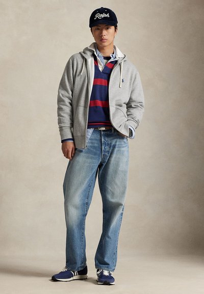 Junge Person trägt eine marineblaue "Ralph"-Kappe, einen grauen Kapuzenpullover mit Reißverschluss, ein gestreiftes marineblau-rotes Rugbyshirt, hellblaue Jeans und blaue Sneakers, steht lässig.