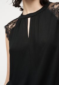 Μαύρο αμάνικο μπούστο με μανίκια από δαντέλα και μικρό άνοιγμα με σχήμα κλειδαρότρυπας κάτω από το στρογγυλό ντεκολτέ, φορεμένο από άτομο με κοντά σκουρόχρωμα μαλλιά.