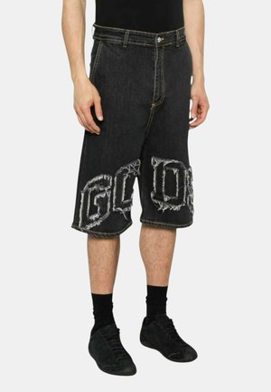 Uomo che indossa pantaloncini di jeans neri al ginocchio con grandi lettere sfrangiate e sneakers nere con calzini, in piedi su uno sfondo semplice.