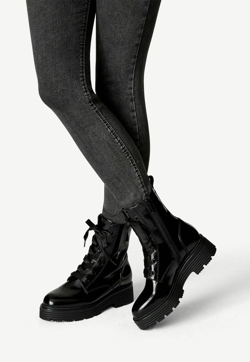 Jambes en jean skinny gris foncé portant des bottines noires brillantes à lacets avec des semelles épaisses en caoutchouc, debout, croisées sur un fond blanc uni.