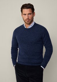 Maglione lavorato a maglia blu navy con scollo rotondo e polsini a coste. Texture liscia con un motivo sottile. Abbinato a una camicia celeste con colletto.
