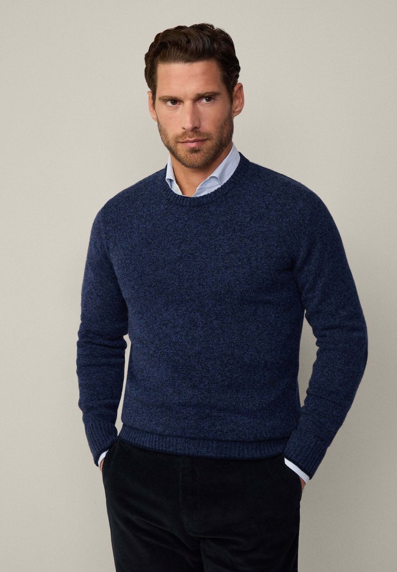 Maglione lavorato a maglia blu navy con scollo rotondo e polsini a coste. Texture liscia con un motivo sottile. Abbinato a una camicia celeste con colletto.