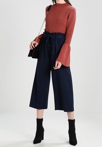 Rostfärgad ribbad långärmad topp med utsvängda ärmar, i kombination med marinblå vida byxor med bälte i midjan och svarta ankelstövlar.