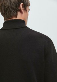 Giovane uomo con capelli corti castani si allontana, indossando un maglione a collo alto a coste nero su uno sfondo chiaro e semplice.