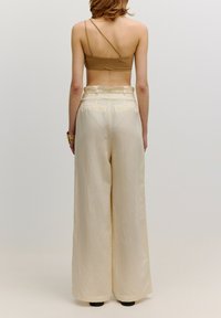 Beige wijde broek met hoge taille, met een gladde textuur en subtiele glans. Achteraan zijn riemlussen en twee achterzakken zichtbaar.