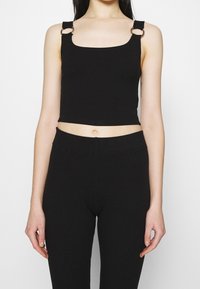 Schwarzes, geripptes Crop-Top mit quadratischem Ausschnitt und verstellbaren Trägern, versehen mit runden Metallakzenten. Kombiniert mit passenden, hoch taillierten schwarzen Leggings.