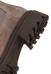 Botte en suède marron avec un dessus en tissu texturé, semelle en caoutchouc marron robuste dotée de crampons profonds pour la traction, et des détails cousus le long des coutures.