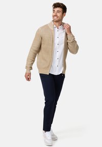 Cárdigan de punto beige claro con cremallera, usado sobre una camisa blanca de botones y combinado con pantalones azul marino y zapatillas blancas. Atuendo informal.