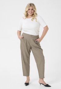 Brown, tapered trousers with a button closure, side pockets, and a subtle texture; paired with a fitted white top and black heeled shoes.