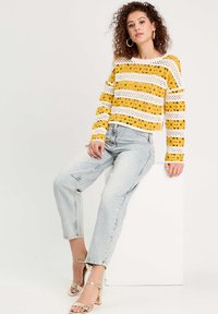 Pull court à rayures jaunes et blanches avec un motif en dentelle, associé à un jean taille haute bleu clair et des sandales à talons blocs métalliques.