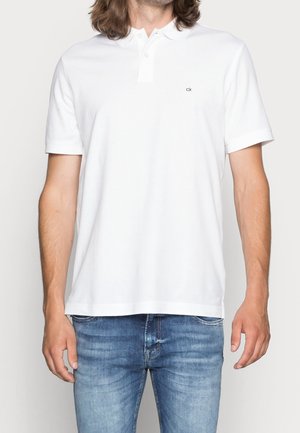Polo blanc en coton, avec une patte de boutonnage à deux boutons, manches courtes et un petit logo sur la poitrine. Texture lisse, coupe standard.