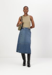 Denim maxi-skjørt med kontraststing, kombinert med en beige tank top. Antrekket har knelange svarte støvler og en grønn håndveske.