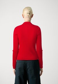 Osoba z blond włosami spiętymi w kok, ubrana w dopasowany czerwony, prążkowany top z długimi rękawami i czarne spodnie cargo, stojąca na jednolitym tle.