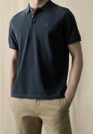 Mand iført en mørkeblå polo med korte ærmer og beige bukser, hænderne i lommerne, foran en neutral baggrund.