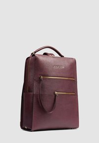Cartella in pelle bordeaux scuro con manico superiore, due tasche frontali con zip e dettagli in metallo dorato su uno sfondo grigio chiaro uniforme.