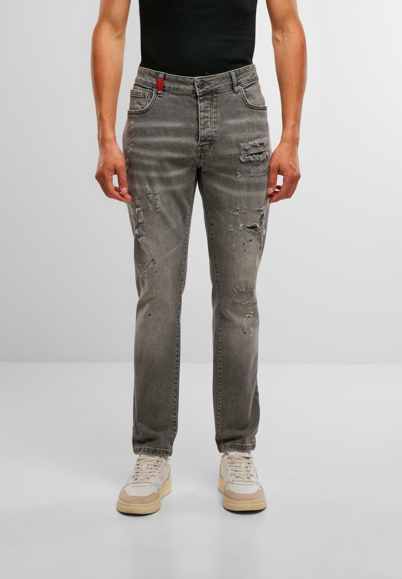 Jeanși din denim gri, cu croială dreaptă, având detalii uzate, un accent roșu la talie și stropi subtili de vopsea albastră.