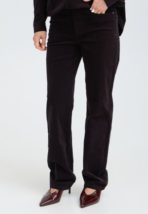 Persona che indossa pantaloni di velluto nero, tacchi scuri a punta e una maglia scura, con una mano in tasca e l'altra lungo il fianco.