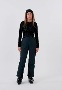 Zwarte top met lange mouwen en een patroonstructuur, gecombineerd met donkerblauwe skibroek met ritsen aan de zijkant en zwarte skischoenen.