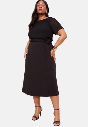 Mujer con vestido midi negro de manga corta, cinturón anudado en la cintura, pulsera dorada de brazalete y sandalias negras de tacón alto, posando con confianza.