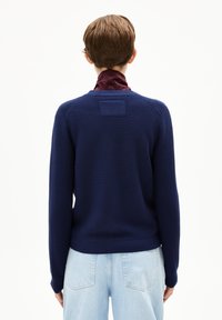 Donkerblauwe gebreide trui met een ronde hals en geribde manchetten, met een textuurontwerp en een vierkant label aan de achterkant. Gecombineerd met lichte spijkerbroek.