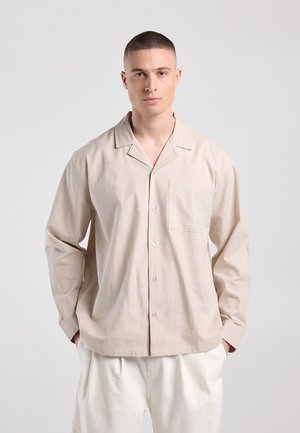 Jonge man met kort haar, gekleed in een beige overhemd met knopen en witte broek, staand met handen in de zakken tegen een effen achtergrond.