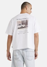 White cotton t-shirt with a relaxed fit, featuring a graphic print on the back, depicting a car and text about balance. Short sleeves.