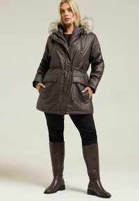 Parka isolée marron avec une capuche en fausse fourrure, taille à cordon de serrage et deux poches à l'avant. Portée avec un pantalon noir et des bottes marron jusqu'aux genoux.