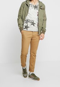 Chaqueta bomber verde oliva sobre una camiseta gráfica gris con estrellas negras, pantalones chinos beige y zapatillas verdes con detalles marrones.