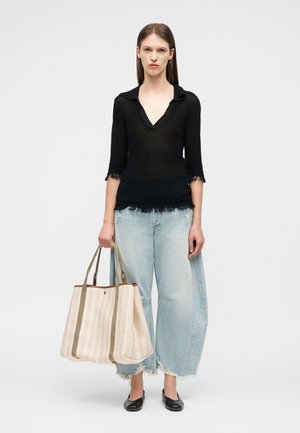 Γυναίκα με μαύρο τοπ σε σχήμα V και φαρδιά ανοιχτό μπλε τζιν κρατάει μεγάλη ριγέ μπεζ τσάντα ώμου, στέκεται μπροστά σε λευκό φόντο.