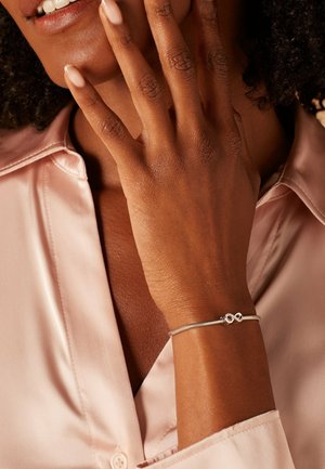 Silberne Armbanduhr mit Unendlichkeit-Design, glatte Textur, getragen an einer Hand mit hellbrauner Haut, vor einem sanften rosa Satinhemd.