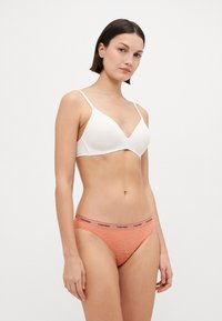 Fehér, sima felületű, vezeték nélküli melltartó, lila csipkés, narancssárga bikini alsóval, fekete logós derékpánttal párosítva.