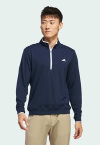 Laine pitkähihaine pullover merivoimansiniseksi, jossa on vale-kaulus, valkoinen vetoketju ja valkoinen yksityiskohta vasemmassa rinnassa, jossa on brändin logo. Sileä kangas rakenne.