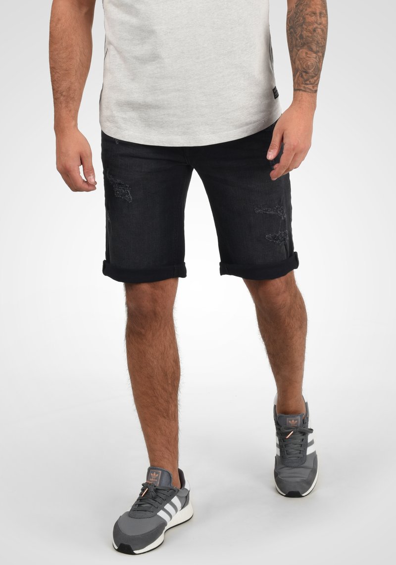 Shorts en denim foncé avec des ourlets retroussés, présentant des détails usés à l'avant. Associés à des chaussures de sport grises et un t-shirt gris clair.