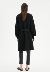 Musta ribbed neuletakki pitkillä hihoilla ja vyöllä, yhdistettynä kiiltäviin mustiin istuviin housuihin ja mustiin liukuviin kenkiin.