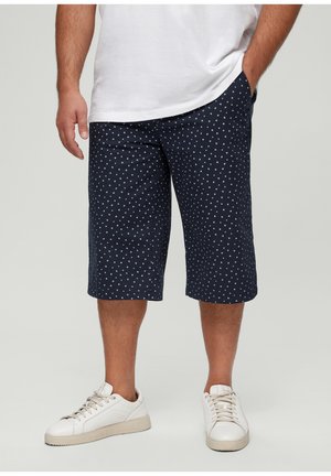 Shorts navy à pois blancs, longueur genoux, en tissu léger. Portés avec un t-shirt blanc et des baskets de couleur claire.
