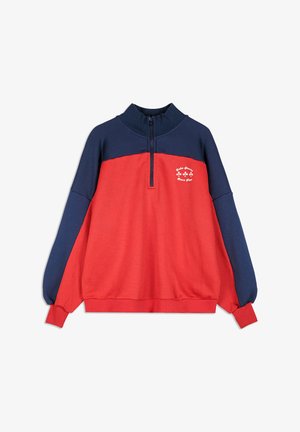 Pull-over à manches longues bleu marine et rouge avec col demi-zip, du petit texte blanc et un graphique de trèfle sur le haut de la poitrine.
